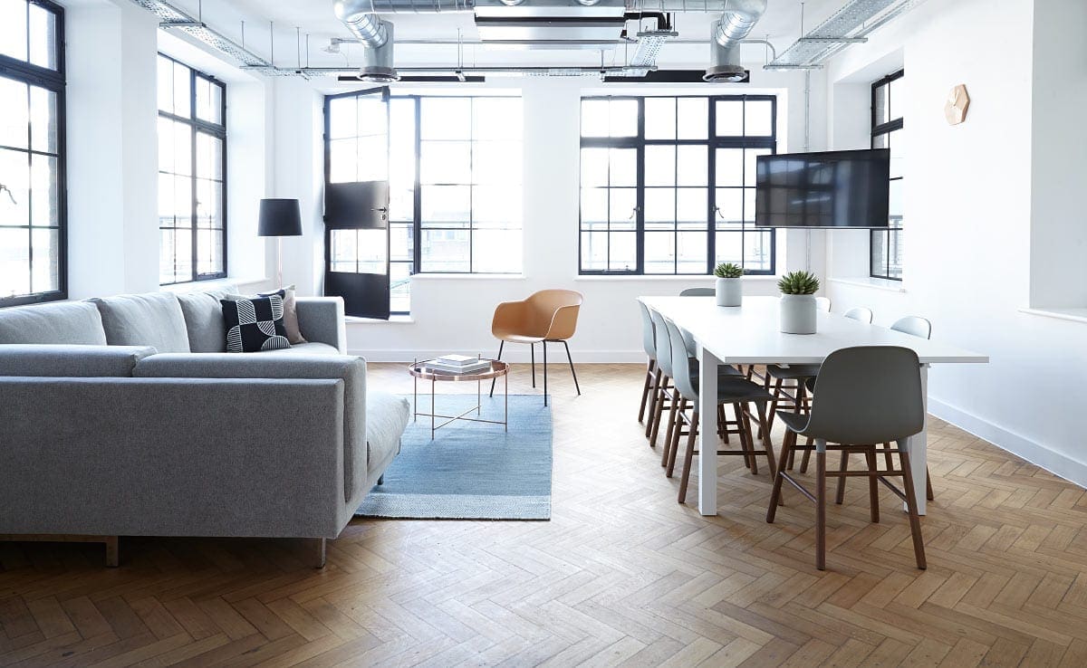 Modern, bright living room with large windows, gray sofa, and dining area with white table and black chairs.