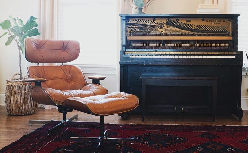 Vintage black upright piano with a brown leather lounge chair nearby in a cozy living room.